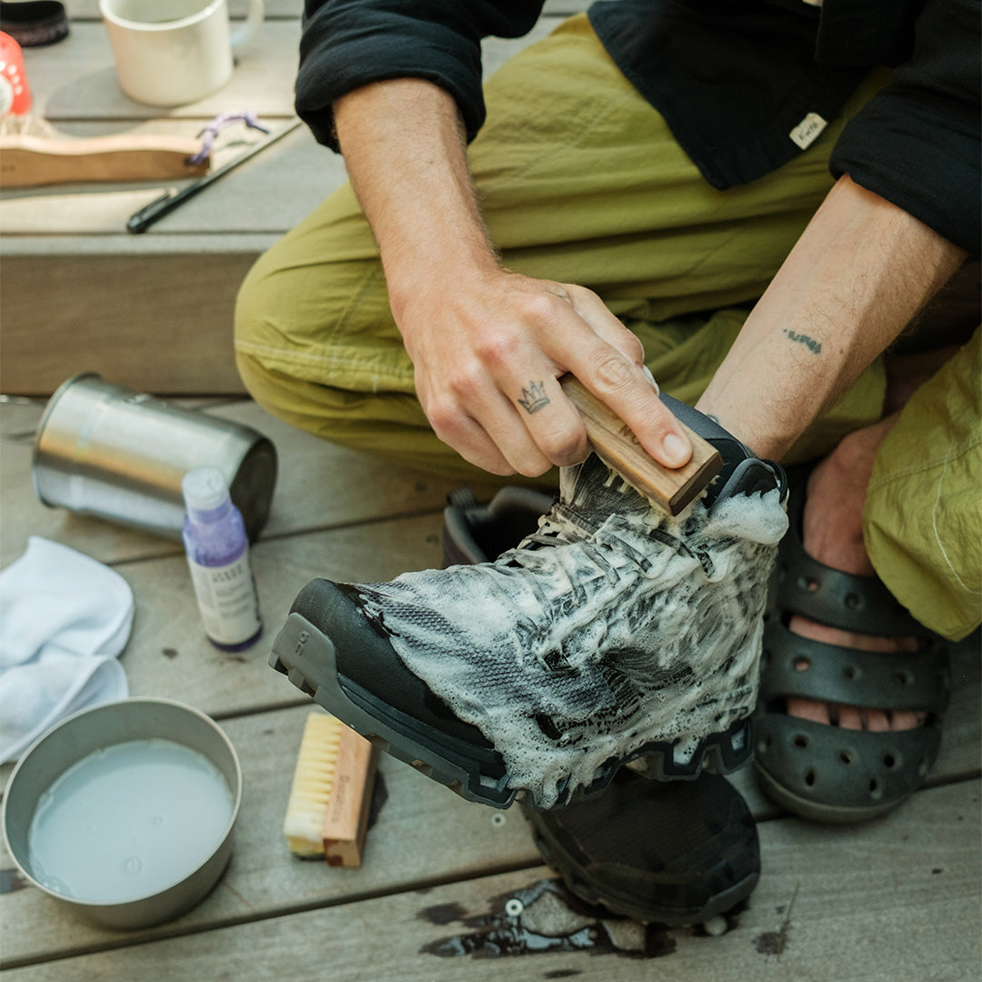 Person cleaning a black boot with a brush on a wooden surface with Deep Cleaning Solution