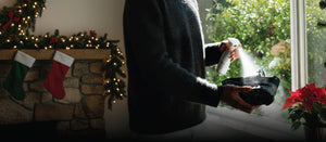 Person spraying shoes with Jason Markk Repel Spray, surrounded by greenery and a potted plant.