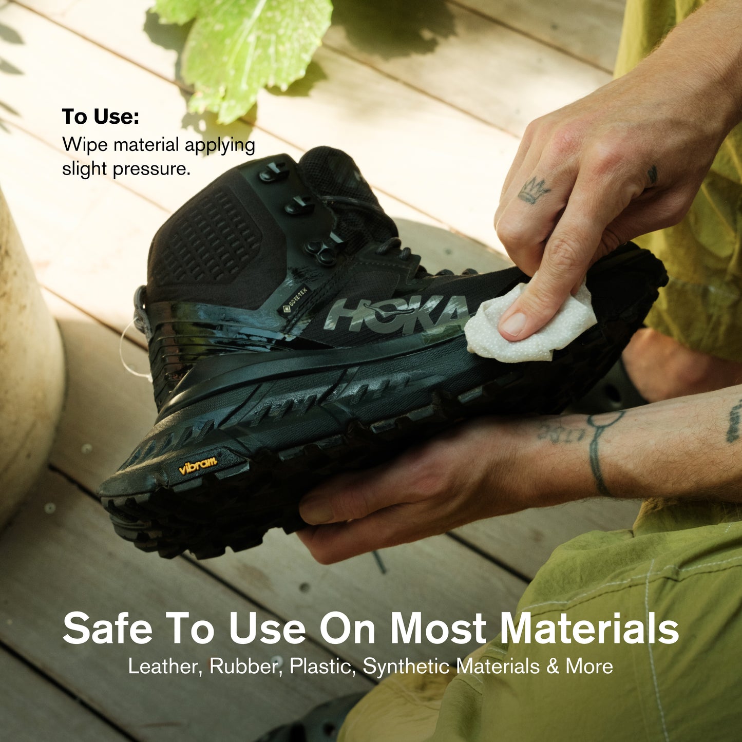A person in olive green pants cleaning a black HOKA GORE-TEX trail hiking boot with a Vibram sole using a Jason Markk shoe cleaning wipe on a wooden deck. Application instructions in the top left read: "To Use: Wipe material applying slight pressure." Text overlay reads: "Safe To Use On Most Materials – Leather, Rubber, Plastic, Synthetic Materials & More."