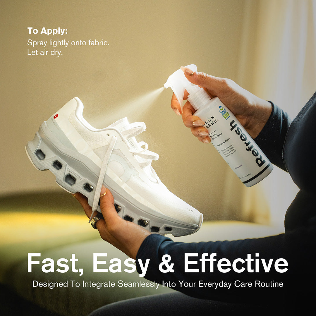 A person in a navy sweater holding a white On Cloudmonster sneaker while spraying Jason Markk Refresh Fabric Spray in Sierra Rain scent directly onto the shoe in a home setting. Application instructions in the top left read: "To Apply: Spray lightly onto fabric. Let air dry." The EPA Safer Choice certified label is visible on the bottle. Text overlay reads: "Fast, Easy & Effective – Designed To Integrate Seamlessly Into Your Everyday Care Routine."