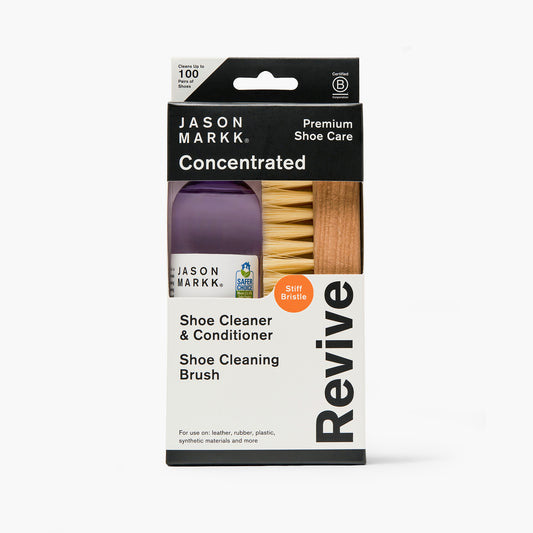 Jason Markk Revive Essential Kit in retail packaging, showing a purple bottle of concentrated shoe cleaner and a wooden stiff bristle brush through a window cutout. Package text: "Jason Markk, Premium Shoe Care, Concentrated, Revive, Shoe Cleaner & Conditioner, Shoe Cleaning Brush, Stiff Bristle, Cleans Up to 100 Pairs of Shoes, For use on: leather, rubber, plastic, synthetic materials and more." Certified B Corporation and EPA Safer Choice seals.