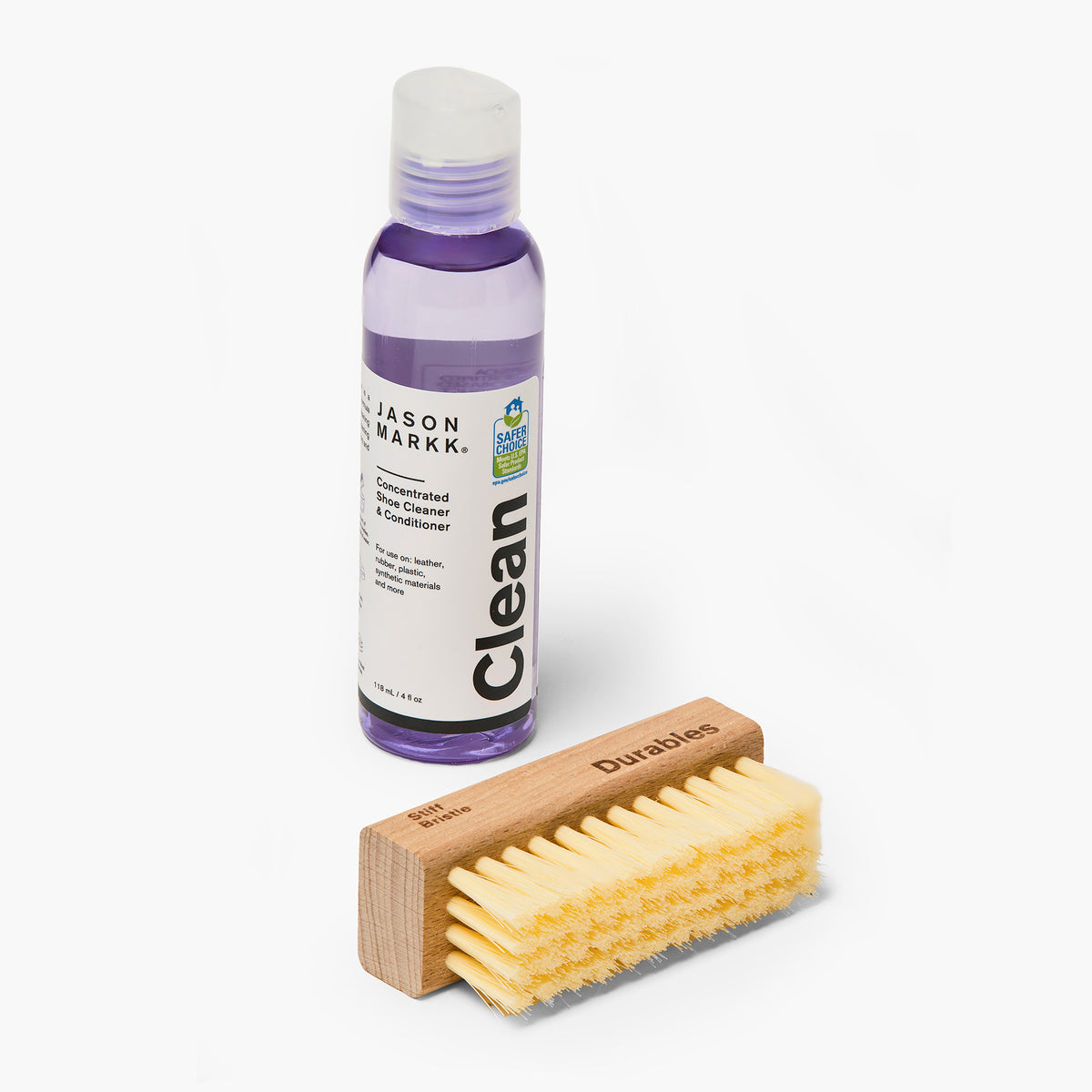 Jason Markk Essential Kit contents out of the box: a clear purple bottle of Concentrated Shoe Cleaner & Conditioner and a wooden stiff bristle brush. Bottle label reads "Jason Markk, Clean, Concentrated Shoe Cleaner & Conditioner, For use on: leather, rubber, plastic, synthetic materials and more, 118 mL / 4 fl oz" with an EPA Safer Choice seal. Brush is engraved "Durables" and "Stiff Bristle."