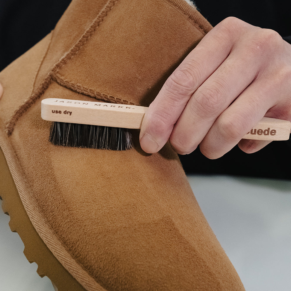 Person cleaning a brown suede Ugg boot with a Suede Brush labeled on a neutral background.