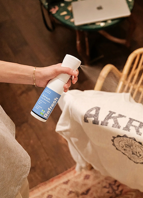 Person spraying Jason Markk Refresh Odor Neutralizer on a white graphic sweatshirt in a home setting. Refresh label reads: Odor Neutralizer, Safer Choice certified. For footwear, hats, and apparel. Demonstrates everyday use of Jason Markk Refresh as a fabric and clothing odor eliminator spray for apparel care and odor control.