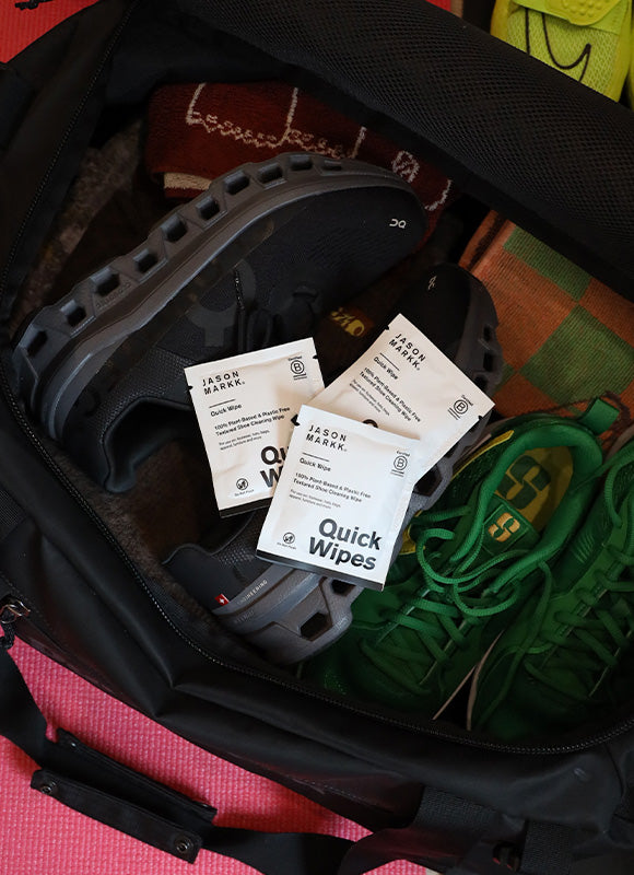 Three Jason Markk Quick Wipes individual shoe cleaning wipe packets inside an open gym bag with sneakers. Quick Wipe label reads: 100% Plant Based and Plastic Free, Textured Shoe Cleaning Wipe. Certified B Corporation. Portable, on-the-go sneaker cleaning wipes for travel, gym bags, and everyday footwear maintenance.