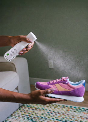 Hand spraying a fine mist from a white spray bottle of Repel onto a purple suede sneaker held in the other hand. The sneaker has a pink upper, a peach side stripe, a blue heel tab, and a white rubber sole. The bottle has a green label and a dark brand name printed near its base. Shown in a living room with a green wall, white armchair, and colorful woven rug, demonstrating a shoe protector spray for suede sneakers.