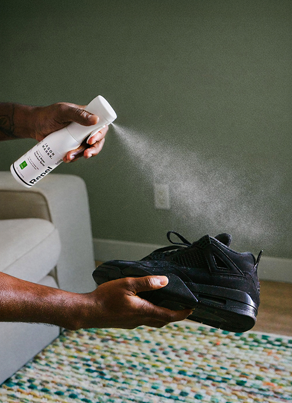 Jason Markk Repel shoe protector spray being applied to black Air Jordan 4 sneakers. A mist of water-repellent spray is visible releasing from the white bottle onto the shoe being held by hand.