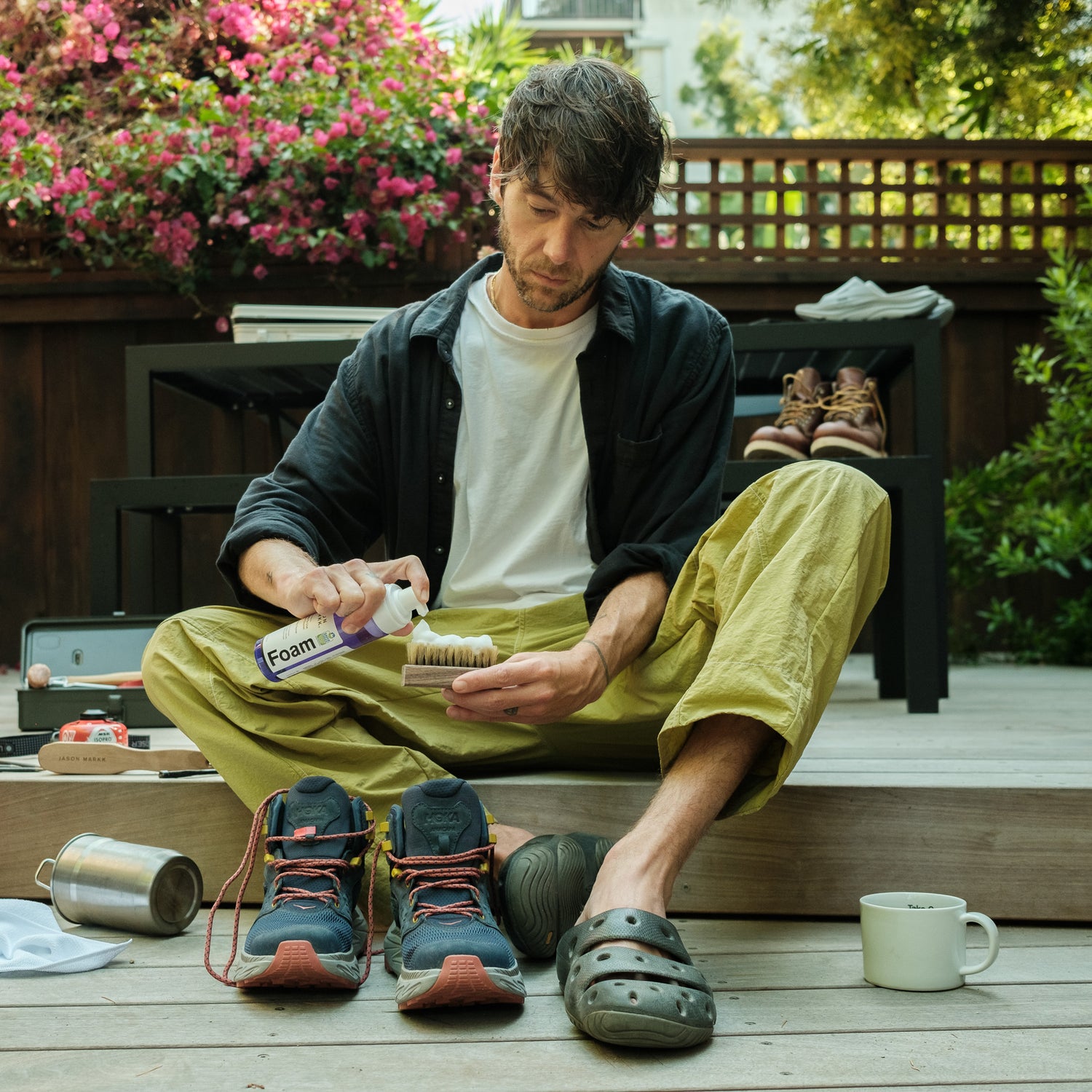 a man sitting on a porch in the backyard cleaning his hoka hiking shoes with jason markk foam cleaner