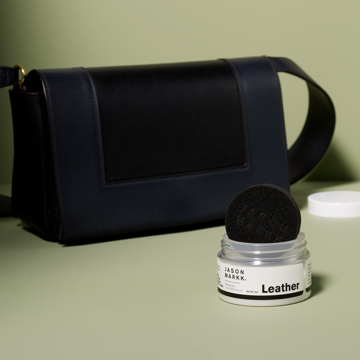 Black leather bag with a jar of Jason Markk leather care balm on a green background