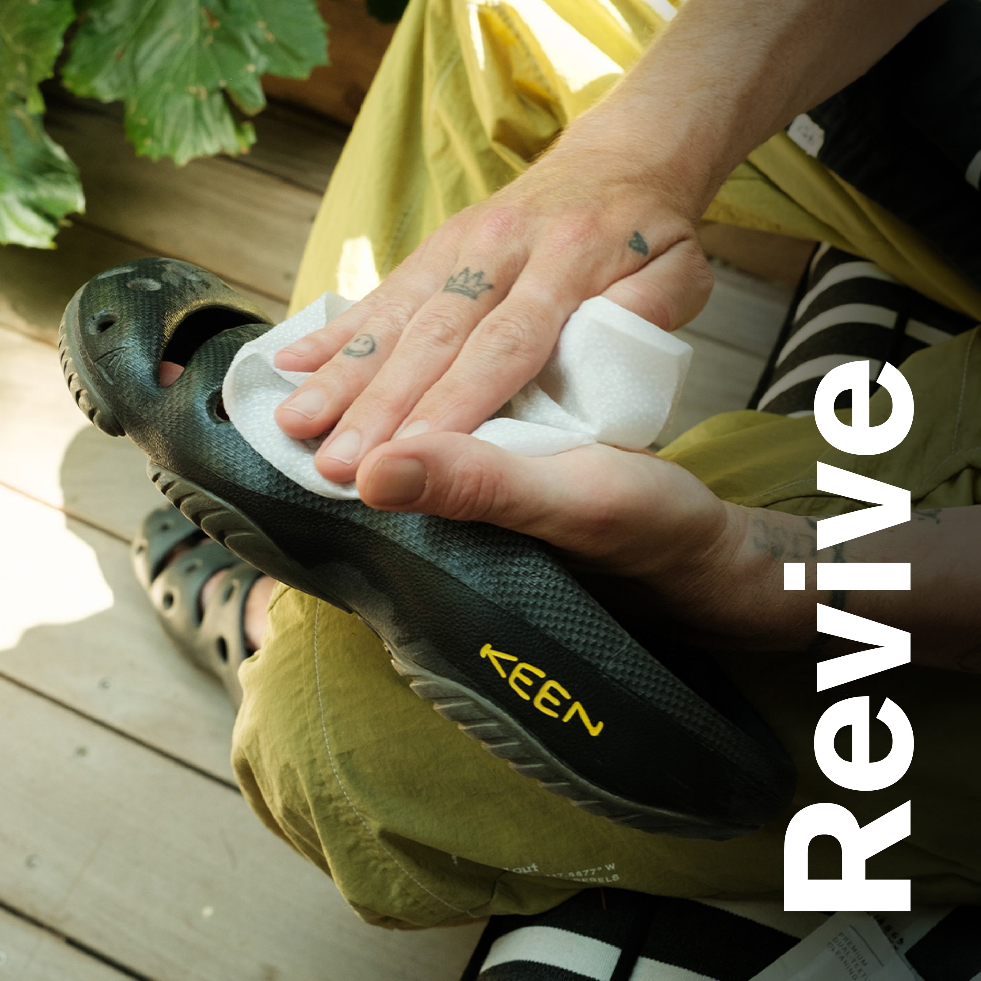 Person wiping a black KEEN clog sandal with a white cleaning wipe, demonstrating shoe care. Visible text includes “KEEN” on the shoe and large vertical overlay text “Revive.” Scene shows hands cleaning footwear outdoors, highlighting quick wipe cleaning for restoring shoes.
