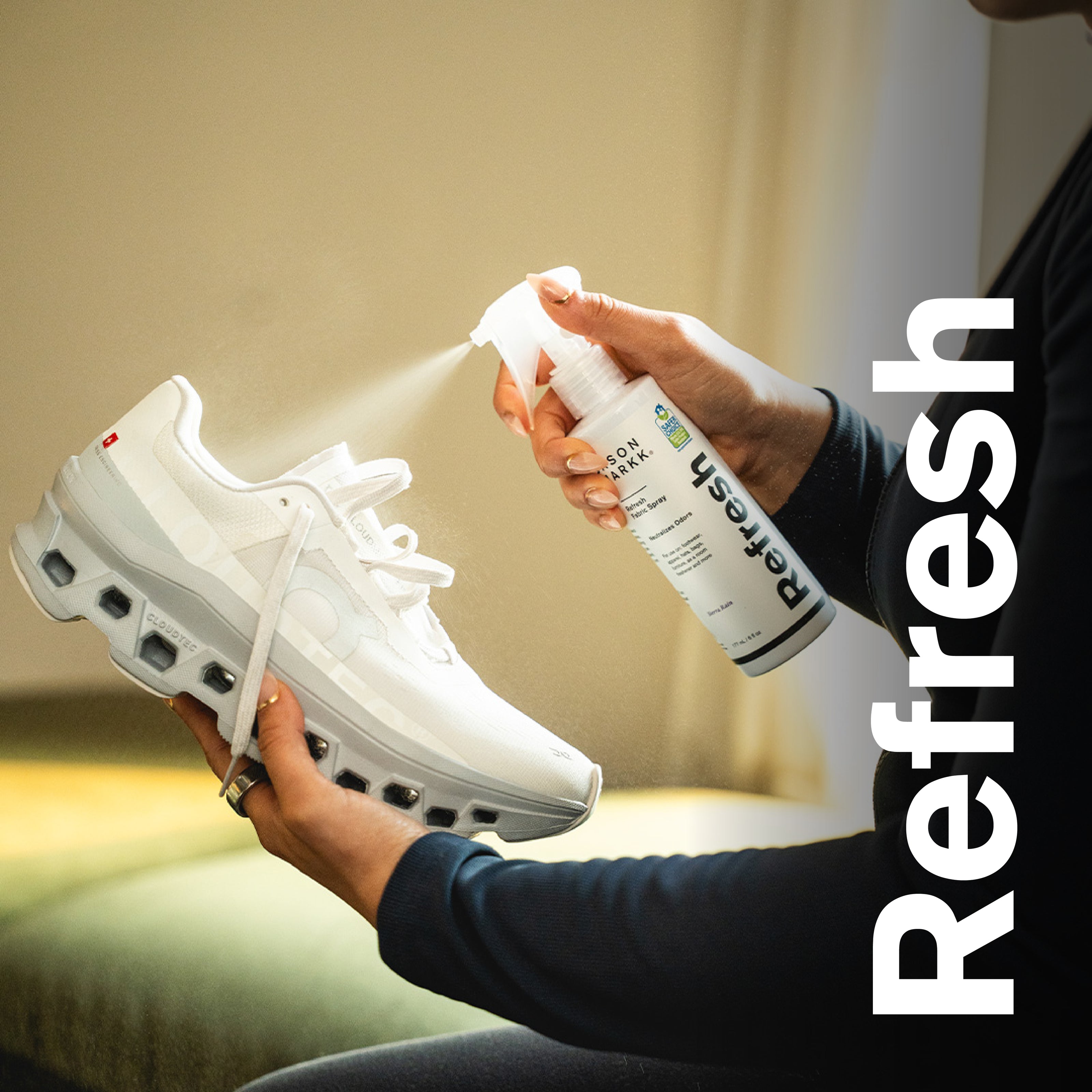 Person spraying Jason Markk Refresh Fabric Spray into a white On Cloudmonster sneaker. The bottle label reads "Refresh Fabric Spray, Neutralizes Odors, EPA Safer Choice Certified, 6 fl oz." Text overlay reads "Refresh." Jason Markk Refresh Spray eliminates odors on footwear, hats, bags, and more.