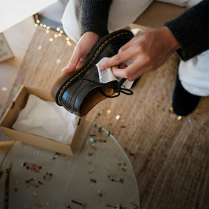 Person cleaning a black leather shoe with a Jason Markk Quick Wipes over a wooden floor.