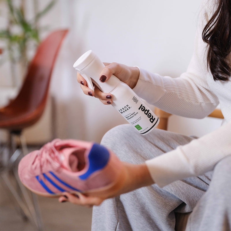Seated person applying Jason Markk Repel Stain & Water Repellent Spray to a pink suede sneaker with blue stripes, in a bright room with a plant and a wood chair. The white bottle reads "Jason Markk, Repel, Stain & Water Repellent Spray, for use on footwear, hats, bags, apparel and more, 159.7 mL / 5.4 fl oz" with a Certified B Corporation seal. No other on-image text.