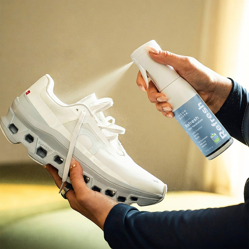 Hands spraying Jason Markk Refresh Odor Neutralizer onto a white running sneaker, with visible mist in warm natural light. The blue and white bottle reads "Jason Markk, Refresh, Odor Neutralizer, Lightly Scented Sierra Rain, for footwear, hats and apparel, 177 mL / 6 fl oz" and carries an EPA Safer Choice seal. No other on-image text.