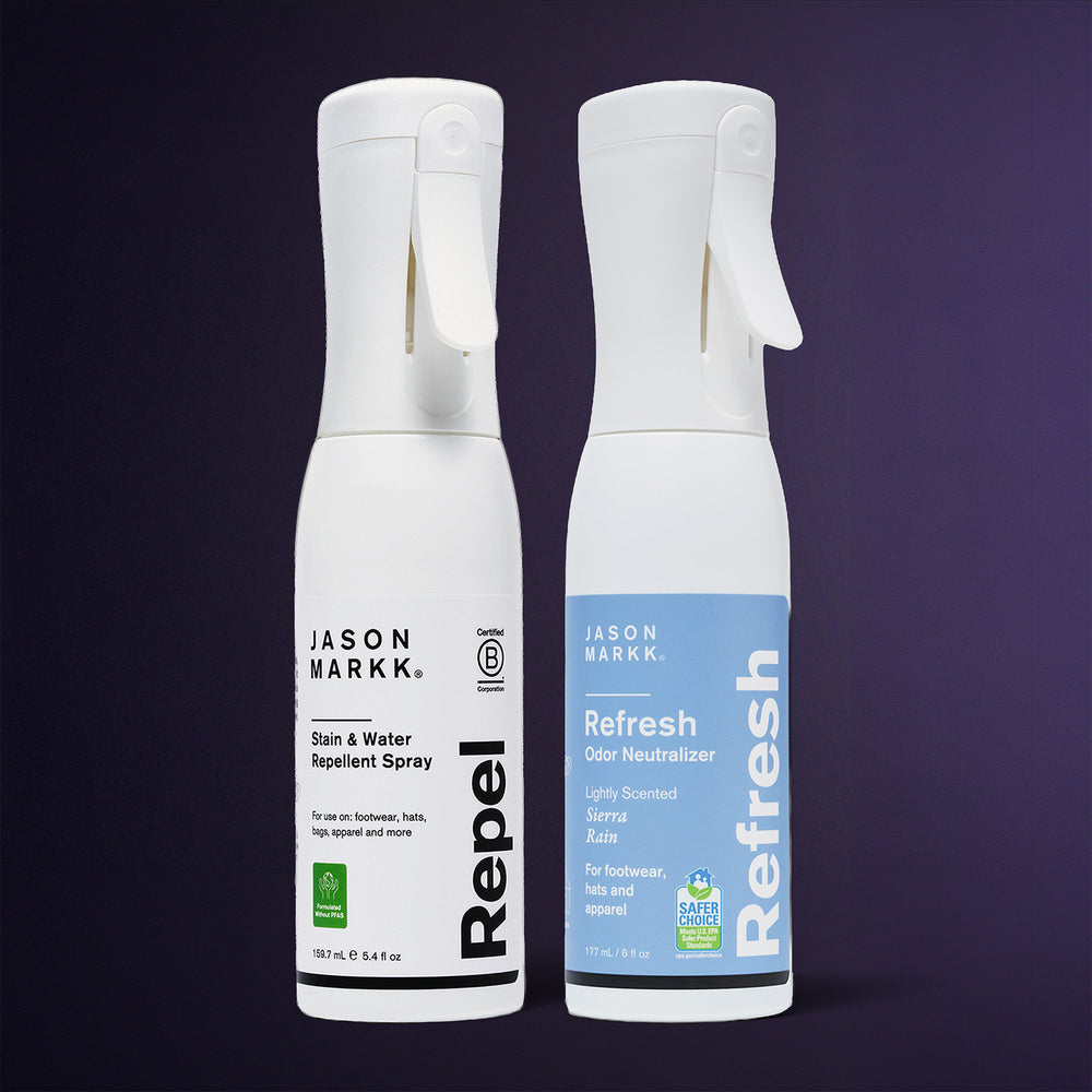 Two Jason Markk spray bottles on a dark purple background. Left: white Repel Stain & Water Repellent Spray, 159.7 mL / 5.4 fl oz, "For use on: footwear, hats, bags, apparel and more," with Certified B Corporation and "Formulated Without PFAS" seals. Right: blue Refresh Odor Neutralizer, lightly scented Sierra Rain, 177 mL / 6 fl oz, "For footwear, hats and apparel," with EPA Safer Choice seal.