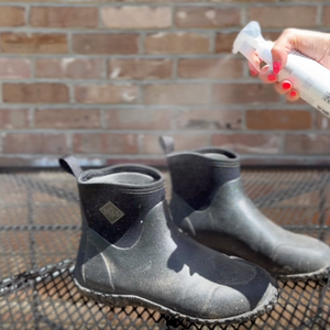 Person spraying black rubber boots with a spray bottle of Refresh against a brick wall.