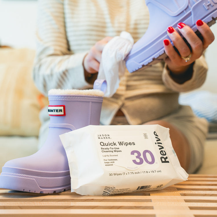 Person in a cream knit sweater wiping a lavender Hunter rain boot with a Jason Markk Quick Wipe. A matching sherpa-lined boot with a "Hunter" logo stands on a wooden slatted table beside a pack of Jason Markk Revive Quick Wipes: Ready-To-Use Cleaning Wipes, Lightly Scented, For footwear, bags and apparel, 30 count, peel & reseal, 30 Wipes (7 x 7.75 inch / 17.8 x 19.7 cm).