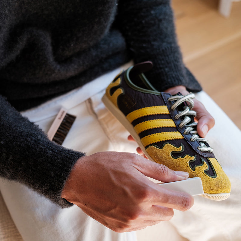 Person holding a yellow and black Adidas sneaker with flame design on a light-colored surface being cleaning with a Premium Suede Kit.