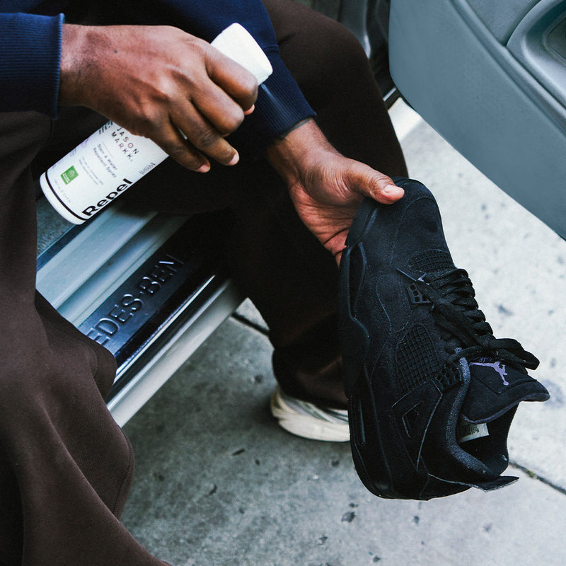Person spraying a black Jordan sneaker with a bottle of Jason Markk Repel Spray on a car step.