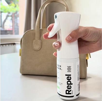 Hand holding Repel with a beige handbag in the background.
